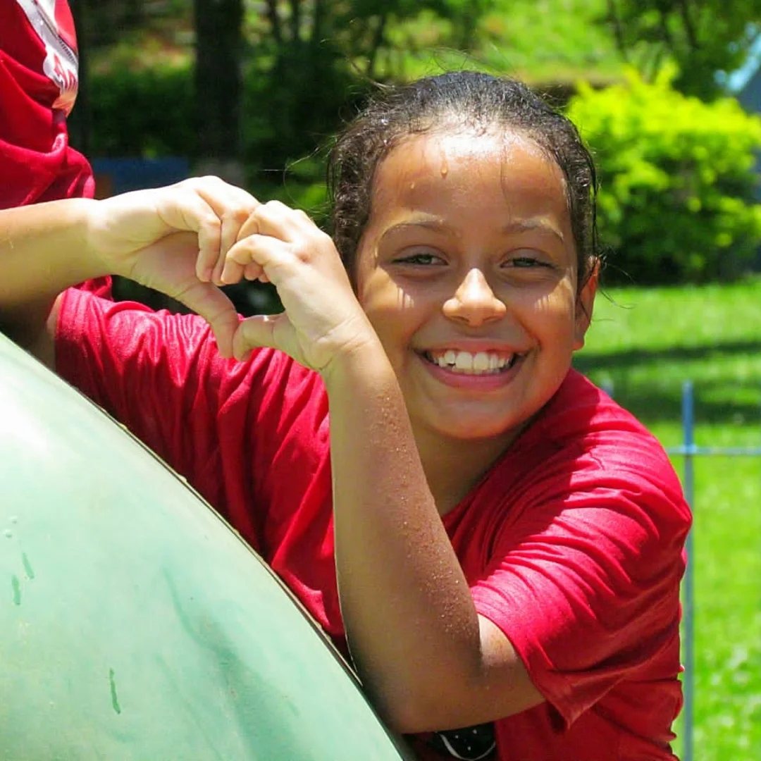 Aluna sorrindo fazendo coração com as mãos durante atividade